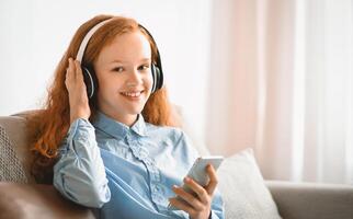 Favorite Song. Smiling Red-Haired Girl Sitting On The Couch At Home, Listening To Music And Touching Wireless Headphones, Holding Her Mobile Phone And Looking At Camera, Enjoying Free Time photo