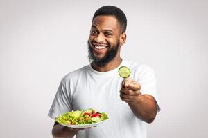 Cheerful Black Man Holding Salad Offering Slice Of Cucumber To Camera Recommending Healthy Eating Standing On White Background. Studio Shot. Try Vegan Diet Concept photo