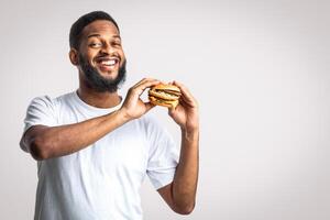 Smiling Black Guy Holding Unhealthy Burger Posing Standing On White Background, Looking At Camera. Male Nutrition And Fast Food Overeating Habit, Cheat Meal On A Diet Concept. Studio Shot photo