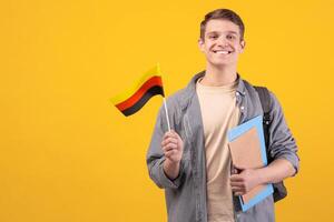 Modern education, language learning and knowledge exchange. Teen cheerful guy in glasses with backpack, notebooks and small Germany flag, isolated on orange background, copy space, studio shot photo