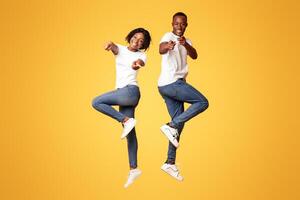 Emotional black couple attractive young man and woman wearing jeans and white t-shirts posing on orange studio background, jumping in the air, pointing fingers at camera. Millennials lifestyle concept photo