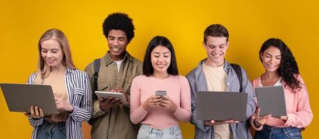 Education, knowledge, modern gadgets in college and social networks. Smiling young multiracial students studying, using digital devices, isolated on orange background, copy space, studio shot photo