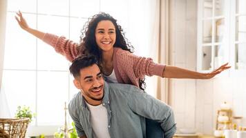Romantic Arab Couple Having Fun Together In Kitchen Interior, Cheerful Middle Eastern Guy Piggybacking His Happy Girlfriend, Joyful Loving Woman Spreading Hands While Having Ride, Copy Space photo