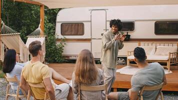 A group of diverse friends relaxes outdoors in a camping setting during summer. They engage in game while seated on a porch near a camper van. video