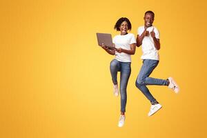 Happy african american millennial man and woman jumping up with laptop, shouting and gesturing on orange studio background. Young black couple gambling online successfully, using laptop, free space photo