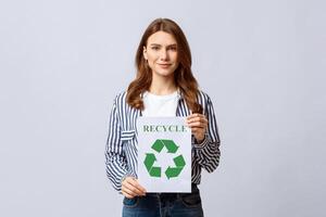 Millennial Eco-Activist Lady Showing Placard With Green Recycle Sign Icon, Promoting Save Environment, Smiling Nature Lover Woman Posing Over Light Studio Background, Looking At Camera, Free Space photo