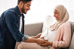 Music For Baby In Womb. Loving Arab Husband Placing Wireless Headphones On Pregnant Wife's Belly While They Resting Together On Couch In Living Room At Home, Muslim Spouses Enjoying Future Parenthood photo
