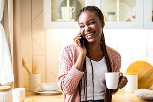 Cheerful black woman talking on mobile phone and drinking coffee in kitchen at home, positive black lady holding mug with hot drink and enjoying pleasant conversation with friend, free space photo