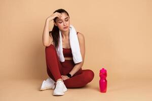 Rest And Relax Concept. Portrait of sporty young woman with white towel on neck sitting on the floor with bottle of water at studio after exercising, taking break. Wellbeing And Training photo