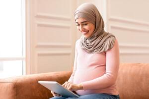 Side View Of Pregnant Muslim Female Using Digital Tablet Computer Browsing Internet Sitting On Sofa At Home. Pregnancy Lifestyle And Modern Gadgets, Technology Concept photo