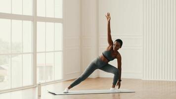 An athletic millennial woman is performing a workout indoors in her home gym. She is focused on maintaining her fitness routine while enjoying the natural light streaming in. video