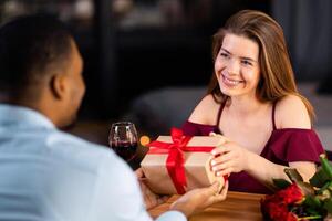 Happy Anniversary. Romantic Man Giving Wrapped Gift Box To His Girlfriend At Resraurant, Greeting With Valentine's Day, Loving Interracial Couple Dining In Elegant Atmosphere, Selective Focus photo