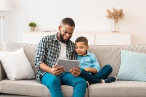 Dad And Son Watching Cartoons On Tablet Online, Sitting Together On Couch Relaxing On Weekend At Home. Happy African American Family Of Two Browsing Internet Using Gadget Computer photo
