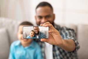 African Father And His Son Making Selfie On Smartphone Bonding And Having Fun Posing Sitting On Sofa At Home. Selective Focus On Selfie Photo On Mobile Phone Screen. Shallow Depth