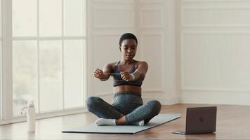 A woman sits cross-legged on a mat while engaging in online workout session at home. She uses a resistance band, focused on maintaining a healthy lifestyle. The room is well-lit with natural light. video