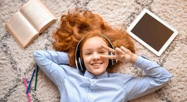 Top above high angle view of positive smiling red-haired teenage girl listening to music, enjoying her favourite song wearing wireless headphones, lying on floor carpet, showing v sign photo