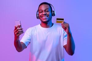 Streaming Music Subscription. Happy black guy in wireless headphones holding smartphone and credit card, paying for audio services online, standing under neon lighting over purple studio background photo