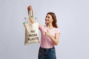 No More Plastic. Smiling Millennial Woman Pointing At Canvas Eco Tote Bag Filled With Empty Bottles Ready For Recycling, Positive Lady Enjoying Zero Waste Living, Standing On Grey Studio Background photo