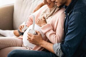 Melodies for Baby In Womb. Expecting muslim couple holding wireless headphones near pregnant belly, turning on music for child development while relaxing together on couch, cropped image, closeup photo