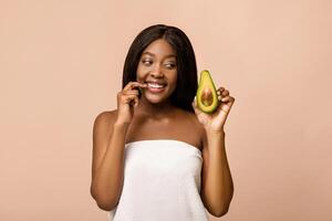Joyful african american lady wrapped in towel holding half of avocado, beige studio background, copy space. Curious black woman looking at avocado, wondering how to use it in face care photo