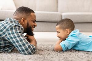 Father's Day. Side View Of Loving African Daddy And Little Son Looking At One Another Lying On Floor Posing At Home. Young Dad And His Kid Boy Bonding Indoor. Father And Child, Family Concept photo