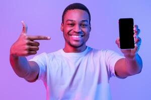 Mobile Offer. Joyful Black Man Pointing At Smartphone With Blank Screen, Indicating Screen Copy Space For App Or Website Design, Standing In Neon Light Over Purple Studio Background, Mockup Image photo