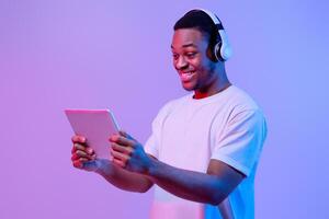 Modern Gadgets. Happy Black Man In Wireless Headset Using Digital Tablet, Playing Online Games Or Watching Videos, Cheerful African Guy Standing In Neon Light Over Purple Background, Copy Space photo