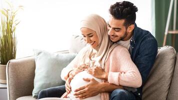 Happy islamic couple expecting baby, hugging on couch at home, cheerful muslim spouses relaxing together on sofa in living room, tenderly touching pregnant belly and smiling, copy space photo