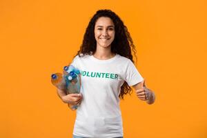 Happy Volunteer Lady Holding Plastic Bottles And Gesturing Thumbs Up To Camera Standing Over Yellow Studio Background. Recycle Your Waste, Keep Environment Clean photo