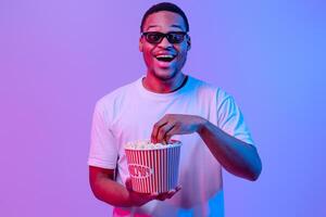 Portrait Of Emotional Black Guy In 3D Glasses With Bucket Of Popcorn, Cheerful African Man Looking At Camera With Excitement, Enjoying Good Movie, Standing Under Neon light Over Purple Background photo
