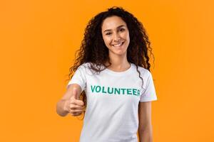 Happy Female Volunteer Gesturing Thumbs Up Approving Charity Work Posing Smiling To Camera Standing In Studio Over Yellow Background. Activist Lady Showing Thumb In Approval. I Like Volunteering. photo