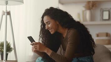 A young Middle Eastern woman relaxes on a cozy couch at home, smiling as she engages with her smartphone. Her casual style reflects a contemporary lifestyle focused on leisure and connectivity. video