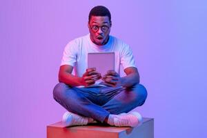 Amazed African Student Guy In Eyeglasses Sitting With Digital Tablet Looking At Screen With Open Mouth, Dazed Black Man Relaxing With Electronic Device On Big Cube Under Neon Purple Light, Copy Space photo