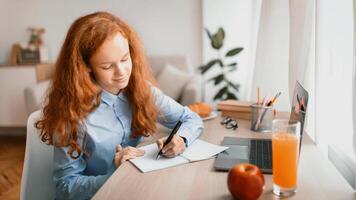 Distance Education Concept. Smiling young teenage girl sitting at desk with laptop and writing in her notebook or diary. Schoolgirl watching online lesson, taking notes, doing homework photo