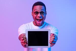 Excited African American Guy Showing Digitral Tablet With Black Screen At Camera, Demonstrating Free Copy Space For Your Website Or App Mockup Design, Standing In Neon Light Over Purple Background photo