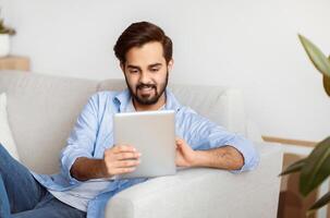 Handsome Eastern Man Browsing Internet On Digital Tablet While Relaxing At Home. Young Smiling Arab Guy Sitting On Couch In Living Room Using Tab Computer, Shopping Online Or Surfing Web, Free Space photo