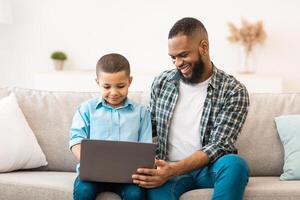 African American Boy And His Father Using Laptop Together, Watching Cartoons Online On Computer Sitting On Sofa At Home. Young Black Dad Bonding With Son Browsing Internet On Gadget Indoor photo