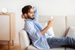 Millennial eastern guy resting on couch with smartphone, browsing internet. Young arab man relaxing with cellphone on sofa in living room, enjoying online communication, side view with free space photo