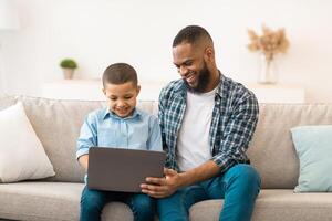 African American Father Teaching Son To Use Laptop Browsing Internet At Home. Little Boy And His Dad Browsing Internet Or Watching Movie Online Together On Weekend. Modern Gadgets Lifestyle photo