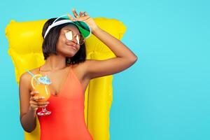 Pretty black lady in swimsuit lying on inflatable lilo and enjoying tropical cocktail over blue studio background. Beautiful African American woman relaxing on pool float, having summer chill photo