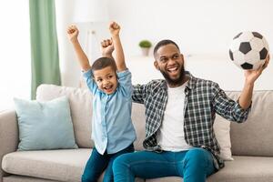 Joyful African Father And Son Watching Soccer Football Game On TV Cheering Favorite Team Sitting At Home. Sport Fans Watch Television. Channel Programming Concept photo