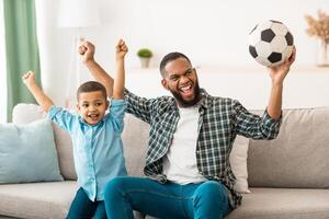 Joyful Black Daddy And Kid Son Watching Soccer Football Game On TV Shaking Fists Cheering Victory Of Favorite Team At Home. Sport Channel Programming Advertisement photo