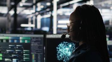 Vertical Data center employee uses audio headset and visual programming dashboard to troubleshoot client reported issues. Server farm technician looks at node tree interface, talking in microphone video
