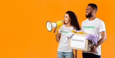 Charity And Volunteering. Two Diverse Volunteers Holding Donation Box Picking Clothes For Homeless People Standing On Yellow Background. Studio Shot, Panorama With Empty Space photo