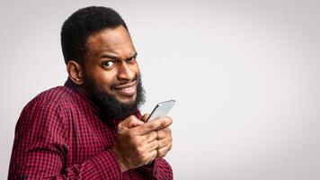 Phone Fraud. Suspicious Black Man Using Smartphone Looking At Camera Posing Over White Studio Background. Telephone Phreaking And Cellphone Scam Concept photo
