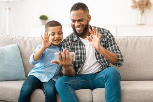 Call. African Father And Son Using Mobile Phone Calling Waving Hands To Smartphone Screen Sitting On Sofa At Home. Remote Online Communication Applications For Cellphone photo
