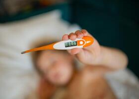 Child holding a digital thermometer. Blurred background with sick boy lying in bed. photo