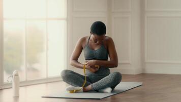 A woman practicing fitness at home sits on a mat, measuring her waistline. She embraces a healthy lifestyle, reflecting on her progress in a bright, inviting space. video
