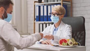 A dietitian consults with a client in a clinic, discussing nutrition and healthy food choices while surrounded by fresh fruits on the table. Both are wearing face masks. video