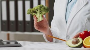 A dietitian in a white coat holds broccoli while explaining dietary choices. Fresh vegetables, including avocado and bell pepper, are laid on the table, cropped video